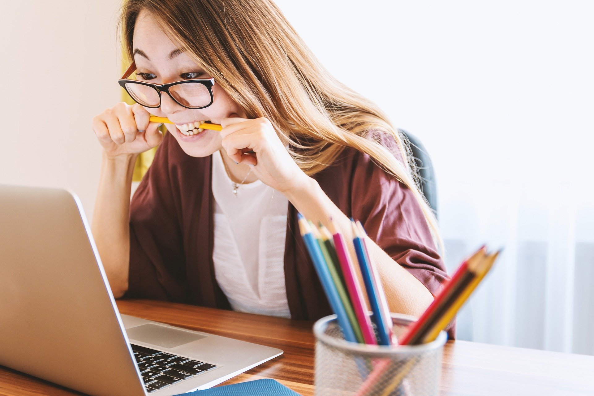 a woman with glasses sits in front of a laptop, chewing on a pencil