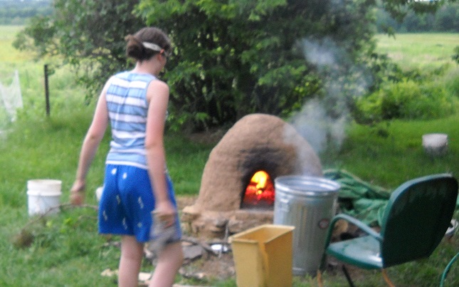 Outdoors in summertime, child wearing swim goggles, shorts, and tank top approaches a domed cob oven with a fire inside it.