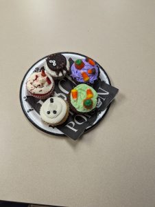 Five cupcakes on a paper plate.  One is white with a ghost face, one is tan, sprinkled with red and with 2 decorations that look like fingers, one is black and has white eyeballs and black insect legs, one is purple and decorated with candy corn and pumpkins, one is green and decorated with candy corn and pumpkins