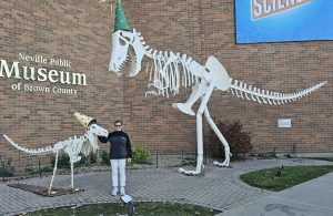 Jennifer Mabie stands next to a small dinosaur skeleton and a large dinosaur skeleton in front of the Brown County Neville Museum