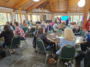 Many people sitting around tables in a big room with many windows